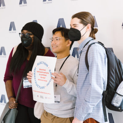 Three people, two wearing masks, pose for a photo holding a sign saying "Election infrastructure is critical to our democracy."