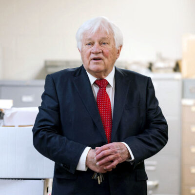 Image of a man wearing a suit and a red tie standing next to a box of papers in a library.